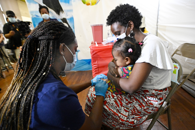 Mother in mask holds daughter on lap while nurse in mask administers shot in arm