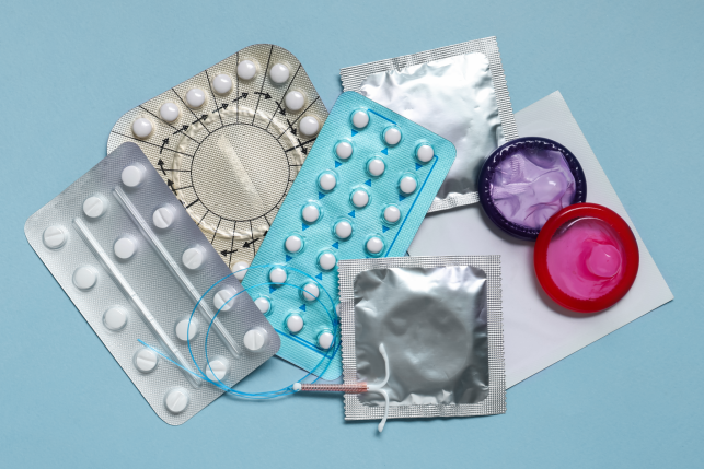 Various packs of contraceptive pills, condoms, and an intrauterine device on a plain background