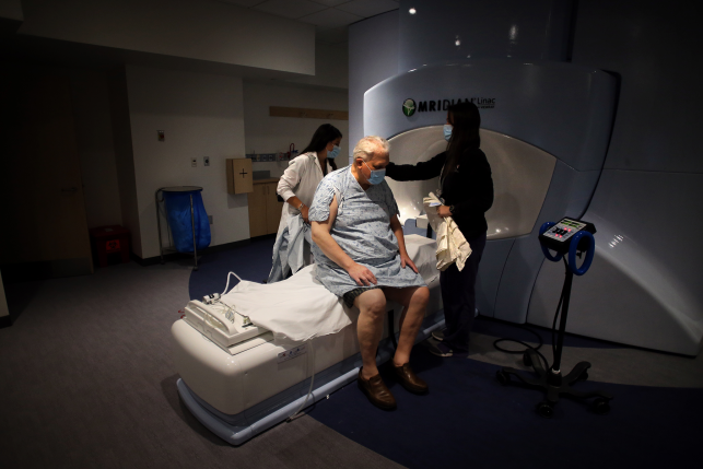 Man in hospital gown and mask waits while nurses prep him for radiology appointment