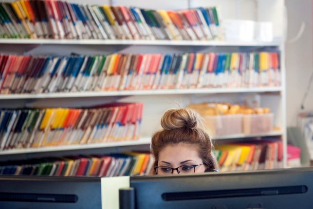 Photo, medical professional speaking on the phone at desk staring at computer monitors with medical records on shelves behind her