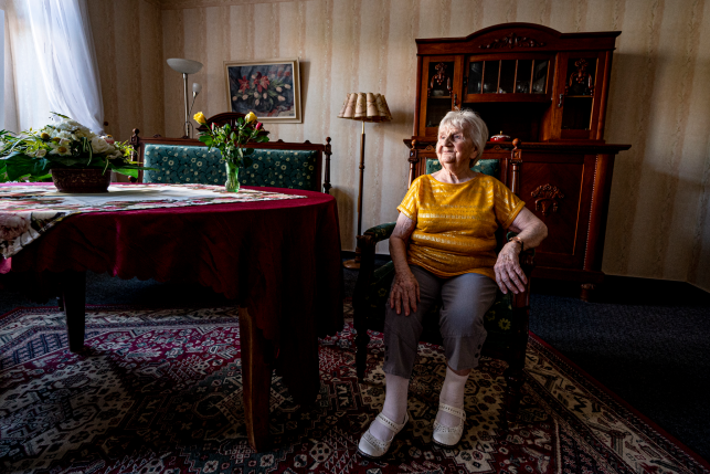 Elderly woman sitting in a chair next to a covered piano smiling and facing the window