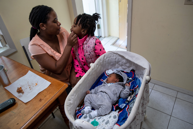 Mother wipes toddler daughters face while baby sleeps next to table in bassinet