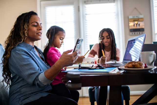 Mom and two daughters sit at dining room table working on computers and school work