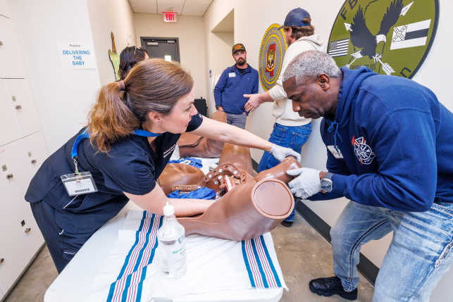 Woman coaching a man through emergency childbirth scenarios with the use of a birthing simulator.