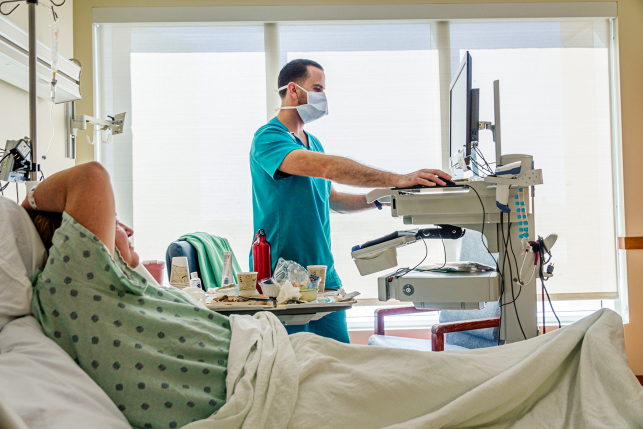 Photo, nurse in mask at computer talking to patient in bed