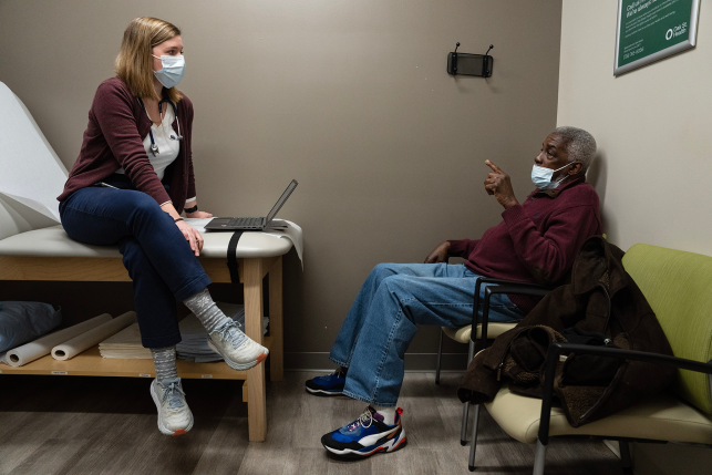 Photo, patient talks to doctor in exam room, both wearing masks.