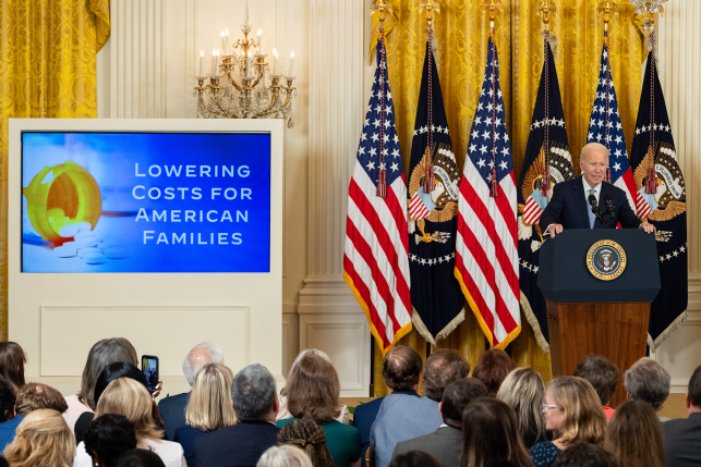 Photo, President Biden at podium in front of flags and powerpoint presentation on lowering costs for American families