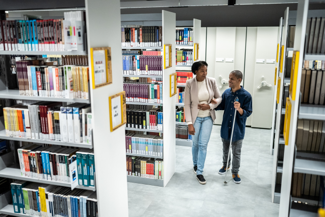 Photo, man with cane walks holding woman's arm in library