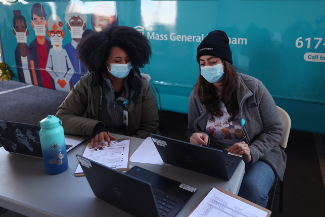 Two women in masks look at computer in front of "Mass General Brigham" sign