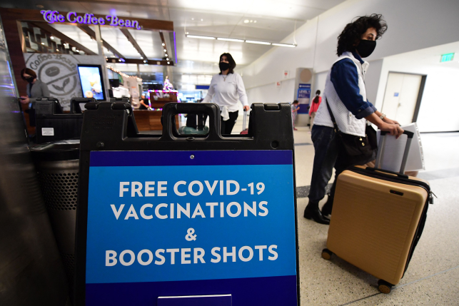 Masked women walk past sign saying "Free COVID-19 Vaccinations & Booster Shots" in brightly lit airport terminal