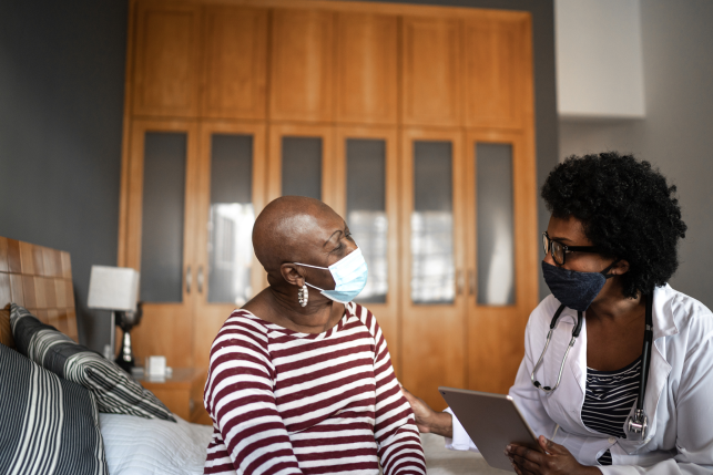 Woman on bed discusses health with her doctor who is holding a clipboard, both wearing masks