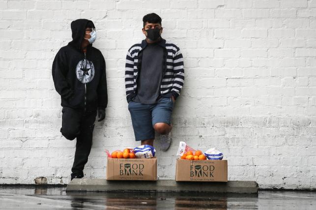 Recipients stand with food they picked up at a food bank in boxes leaning against a wall.