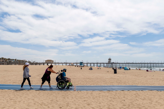 Family pushes boy in wheelchair on mat on the sand of beach with pier in the background