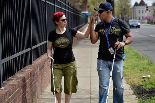 Photo, woman with cane puts fingers up to mouth of man with cane while walking down sidewalk