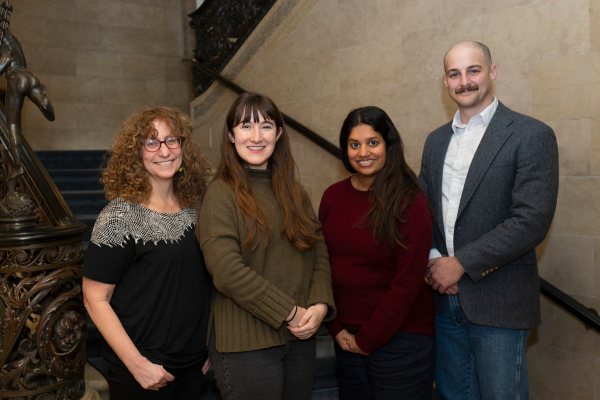 Three women and one man posing for a photo standing in front of a staircase.