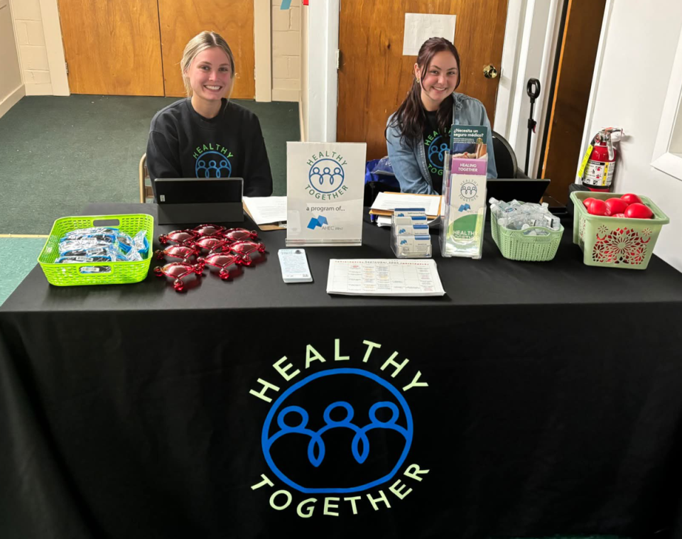 Two women sitting behind a table with a 'Healthy Together' logo tablecloth and various branded giveaways and information