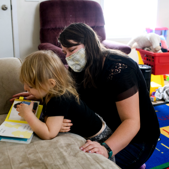 Photo, woman in mask reads to child on couch