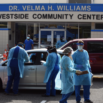 Doctors in scrubs and gowns wait outside cars outside of Dr. Velma H. Williams Westside Community Center