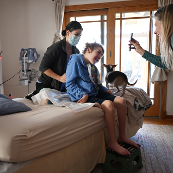 Photo, patient sits on bed facing Mom's phone with nurse at back