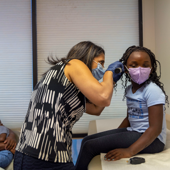 Doctor in mask examines girl in mask's ear on table