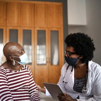 Woman on bed discusses health with her doctor who is holding a clipboard, both wearing masks
