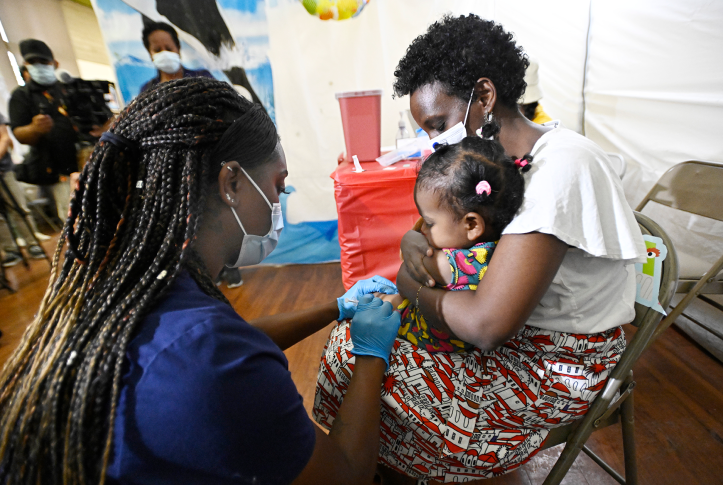 Mother in mask holds daughter on lap while nurse in mask administers shot in arm