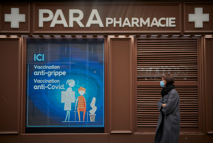 Photo, woman in coat and mask walks past pharmacy signage.