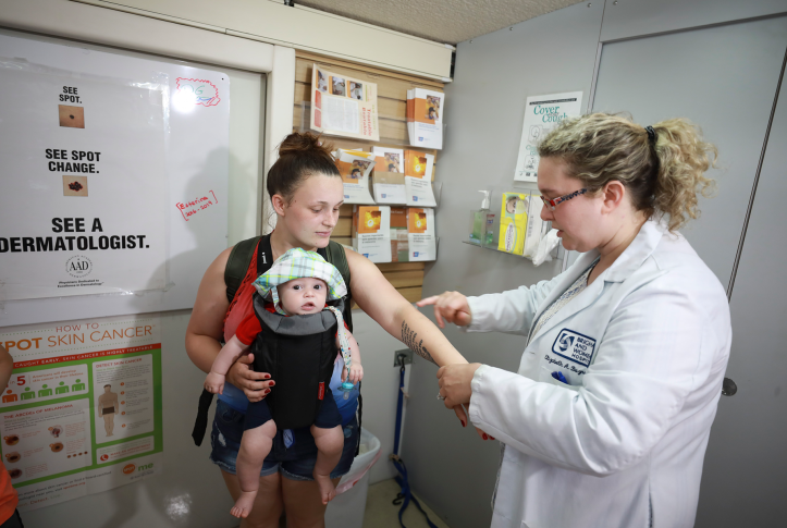 Doctor in white coat examines woman's arm. Woman has baby strapped to chest.