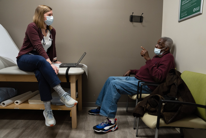 Photo, patient talks to doctor in exam room, both wearing masks.