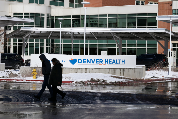 Photo, two people in coats walk in front of the "Denver Health" sign outside a hospital building