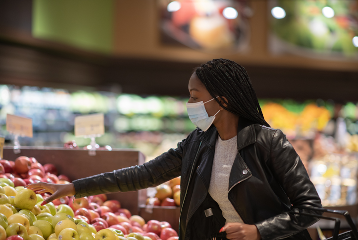 woman in mask reaches to pick an apple from a grocery store shelf