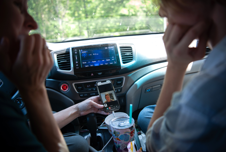 Photo, two people speak on phone in front of car dashboard.