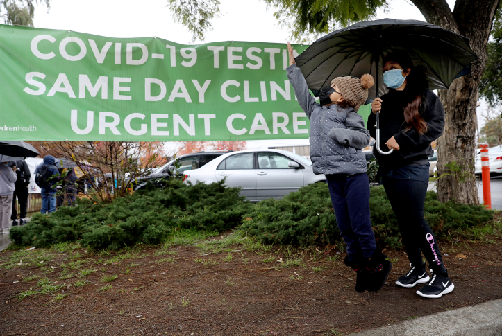 Two young children wait under an umbrella in front of a COVID-19 Testing sign