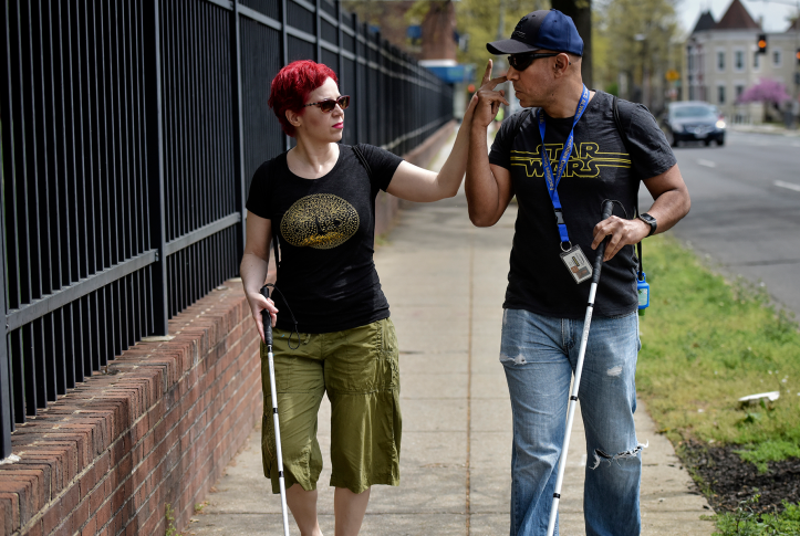 Photo, woman with cane puts fingers up to mouth of man with cane while walking down sidewalk