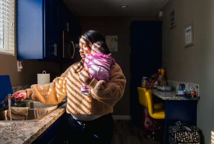 Mom washes cloth in sink while holding newborn baby in her kitchen