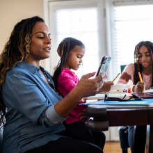 Mom and two daughters sit at dining room table working on computers and school work