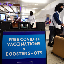 Masked women walk past sign saying "Free COVID-19 Vaccinations & Booster Shots" in brightly lit airport terminal