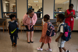 Adult woman bends down to talk to kids entering school with backpacks