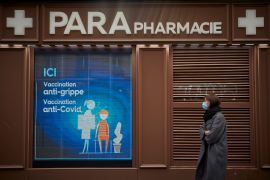 Photo, woman in coat and mask walks past pharmacy signage.
