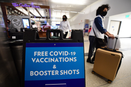 Masked women walk past sign saying "Free COVID-19 Vaccinations & Booster Shots" in brightly lit airport terminal
