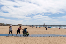 Family pushes boy in wheelchair on mat on the sand of beach with pier in the background